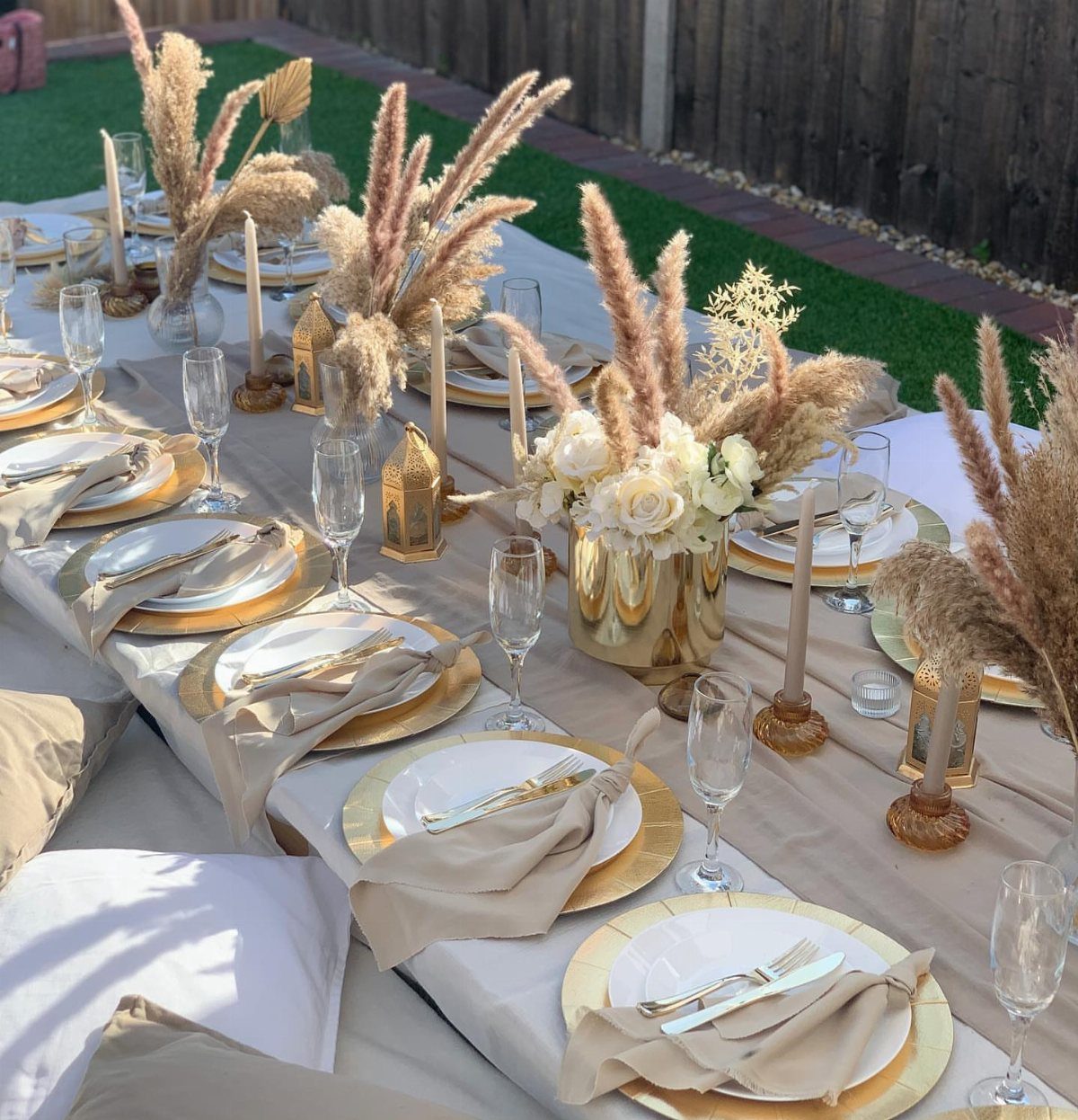 Dining table set with gold vases containing white roses and beige pampas grass.