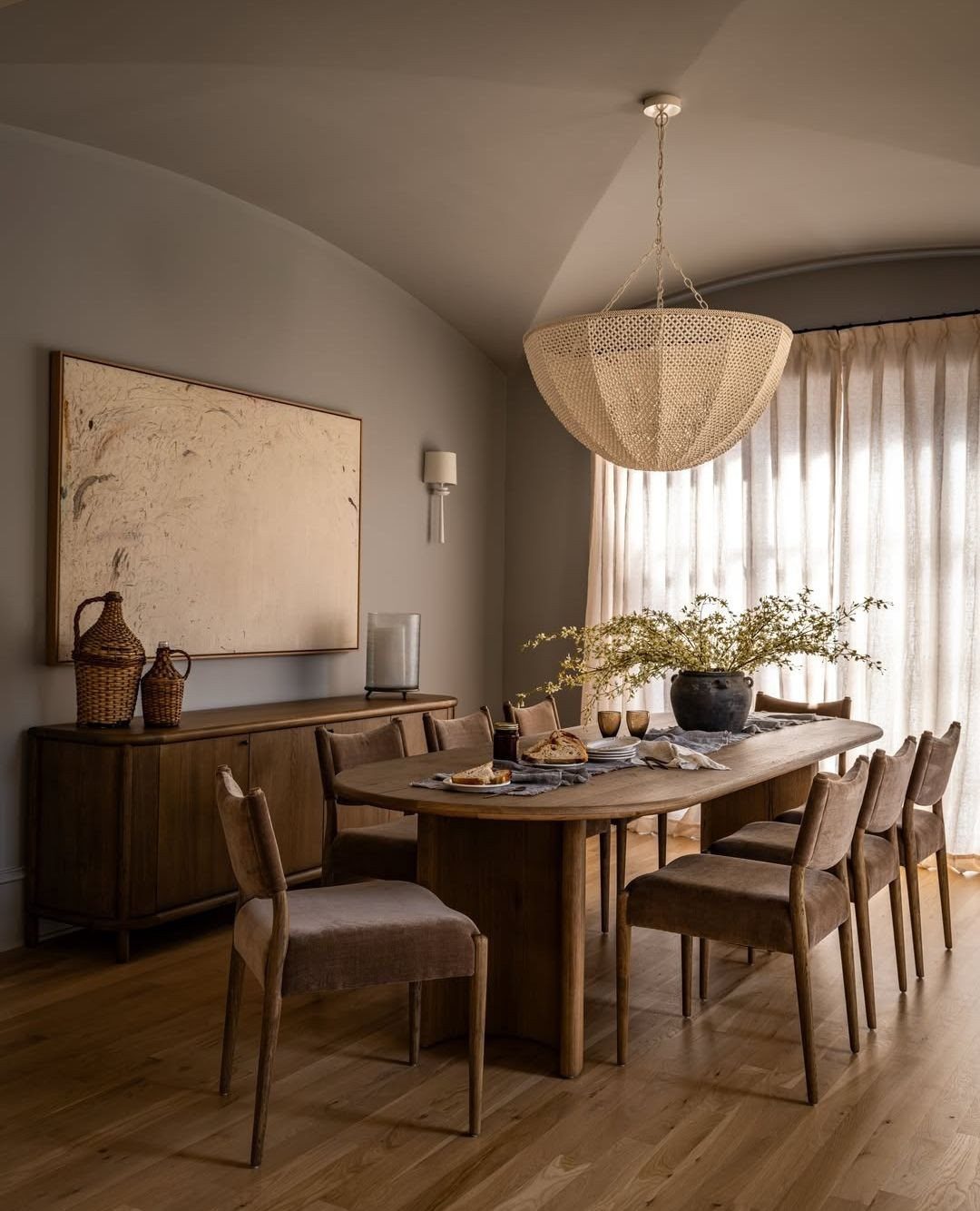 Oval wooden dining table with armless upholstered chairs under a large woven pendant light.