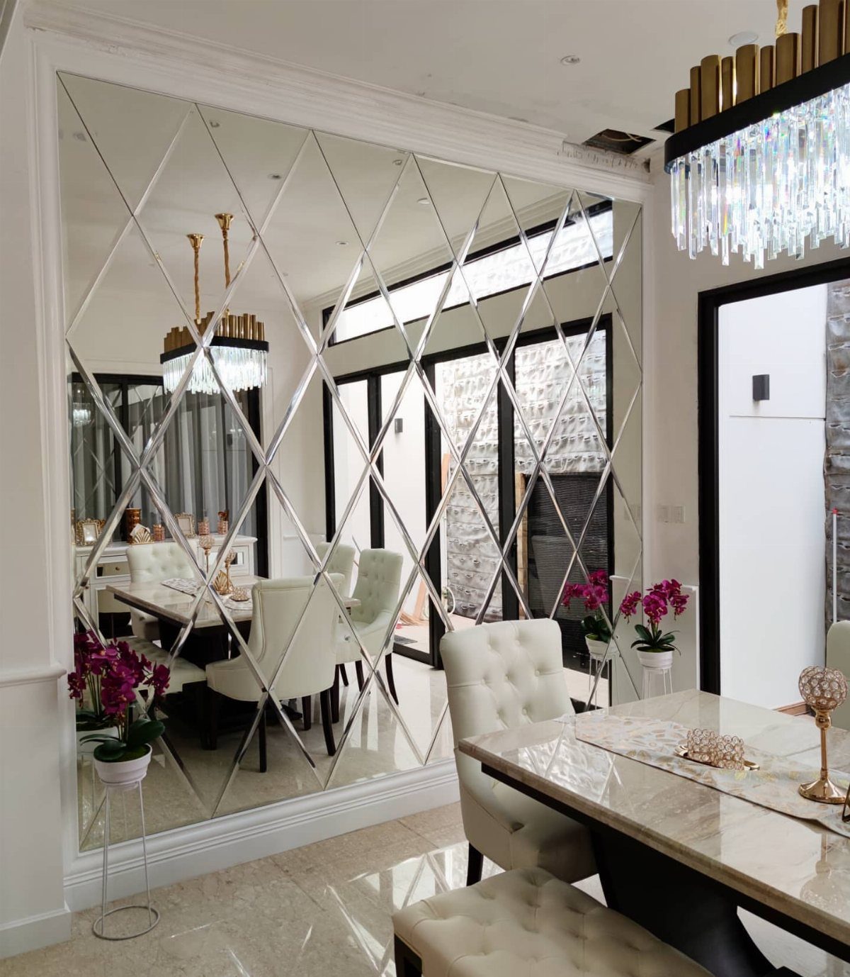 Diamond-patterned mirrored wall reflecting a dining table with white tufted chairs and chandelier.
