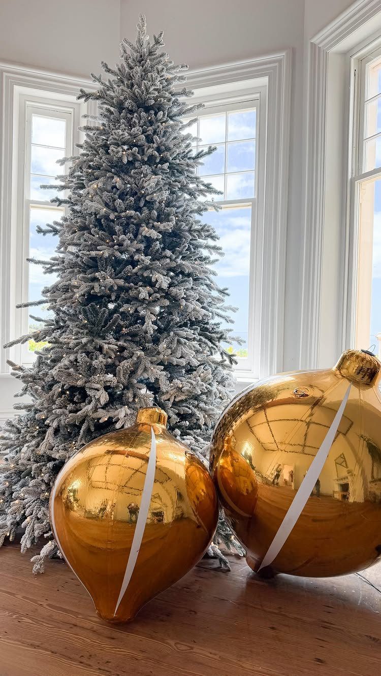 Two oversized gold Christmas ornaments resting on a wooden floor by a flocked tree.