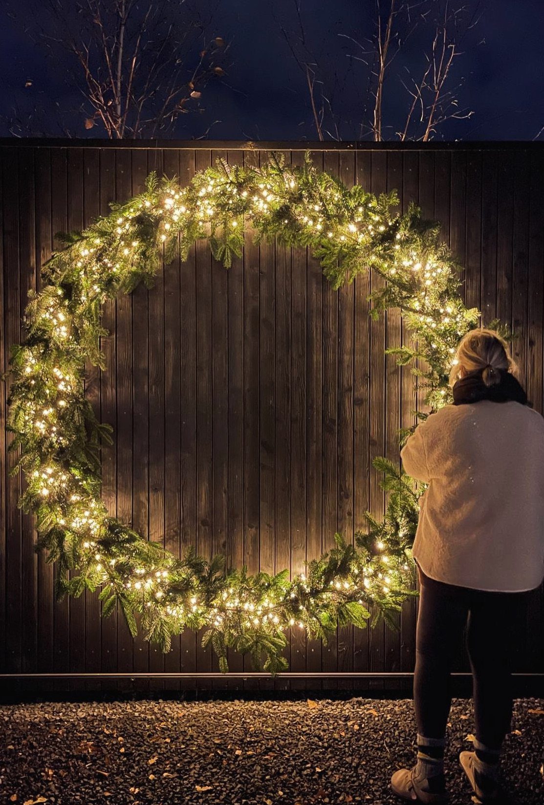 Giant evergreen wreath with glowing fairy lights on a dark wooden fence.