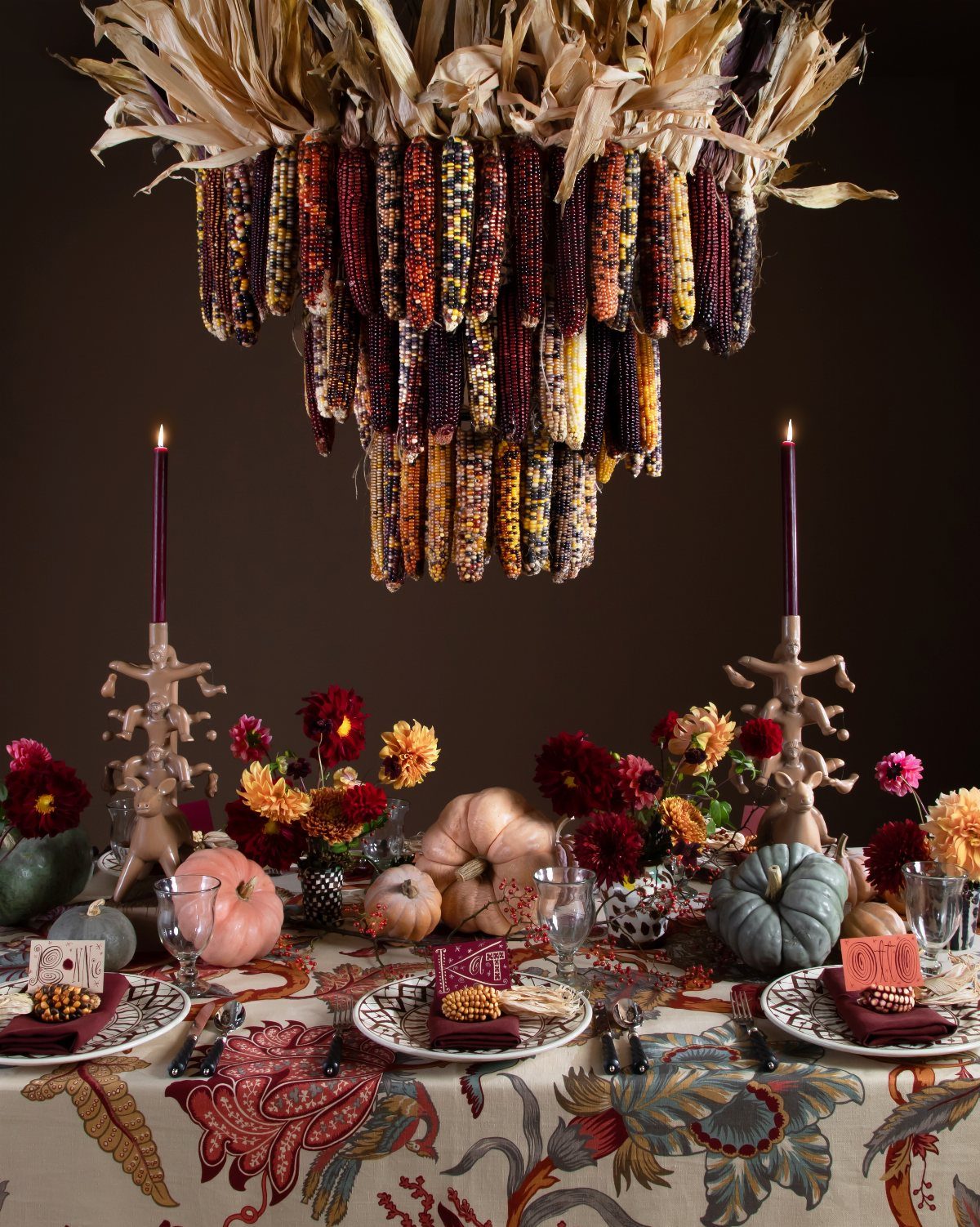 Fall dining table with colorful pumpkins, red flowers, and hanging dried corn installation.