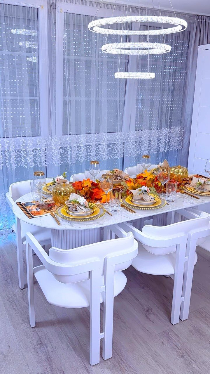 Fall dining table set with gold chargers, autumn leaf garland, and white chairs.