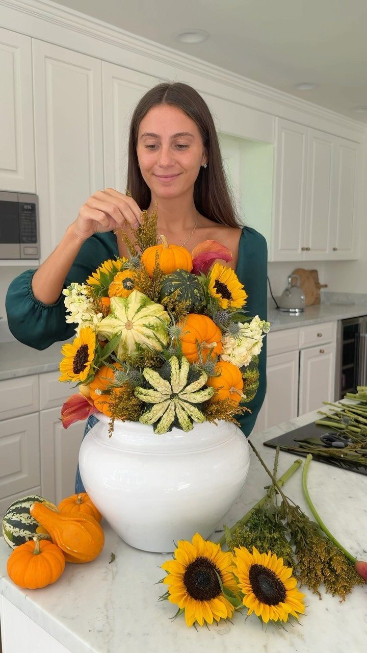 Woman arranging a fall centerpiece with sunflowers and pumpkins in a white bowl.