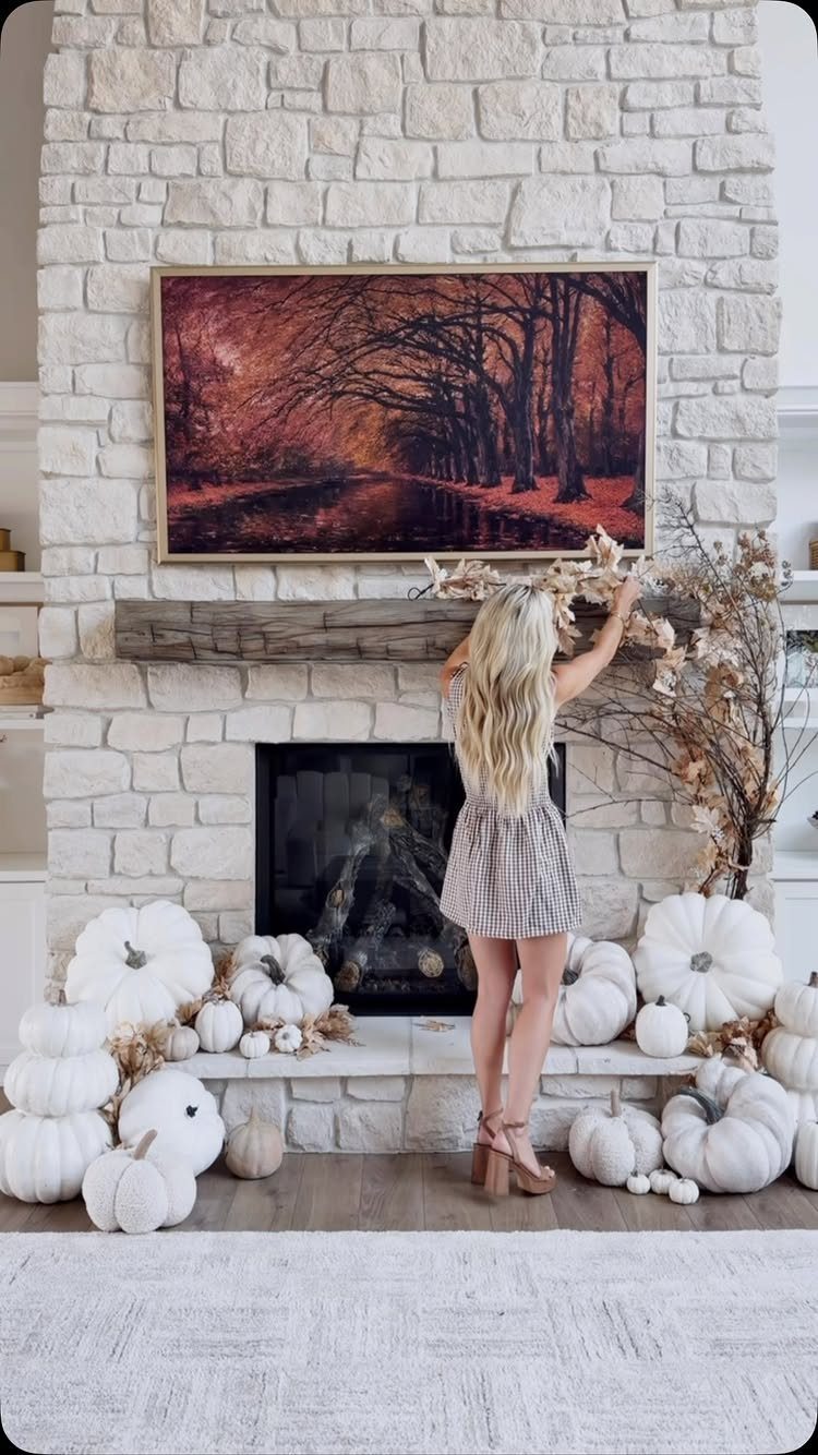 Woman arranging autumn branches on a white stone fireplace mantel with white pumpkins.