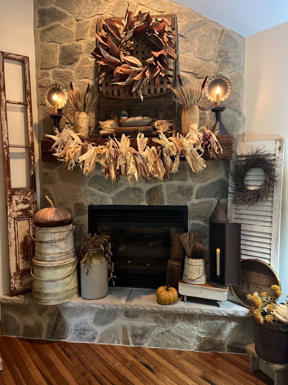 Stone fireplace mantel with draped corn husk garland, lit candlesticks, and autumn pumpkins.