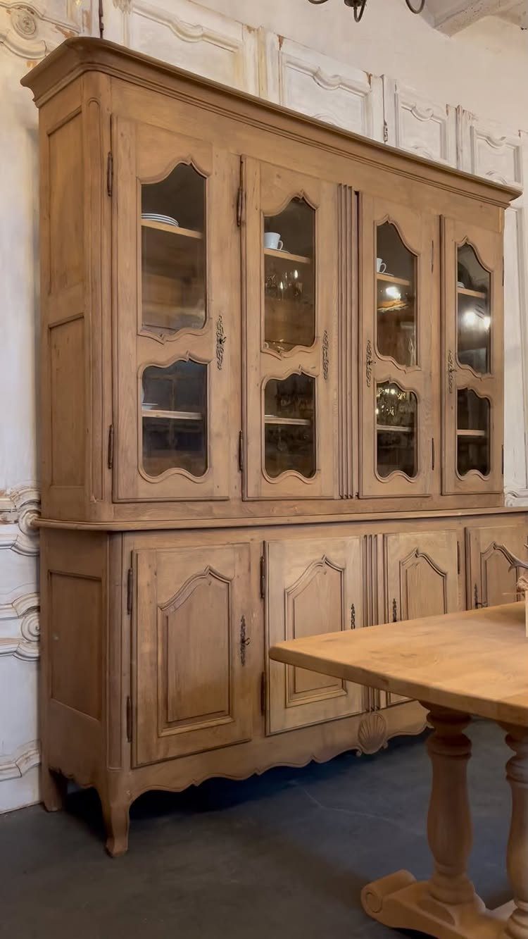 Rustic light-wood hutch with glass cabinets standing against an ornate white wall.