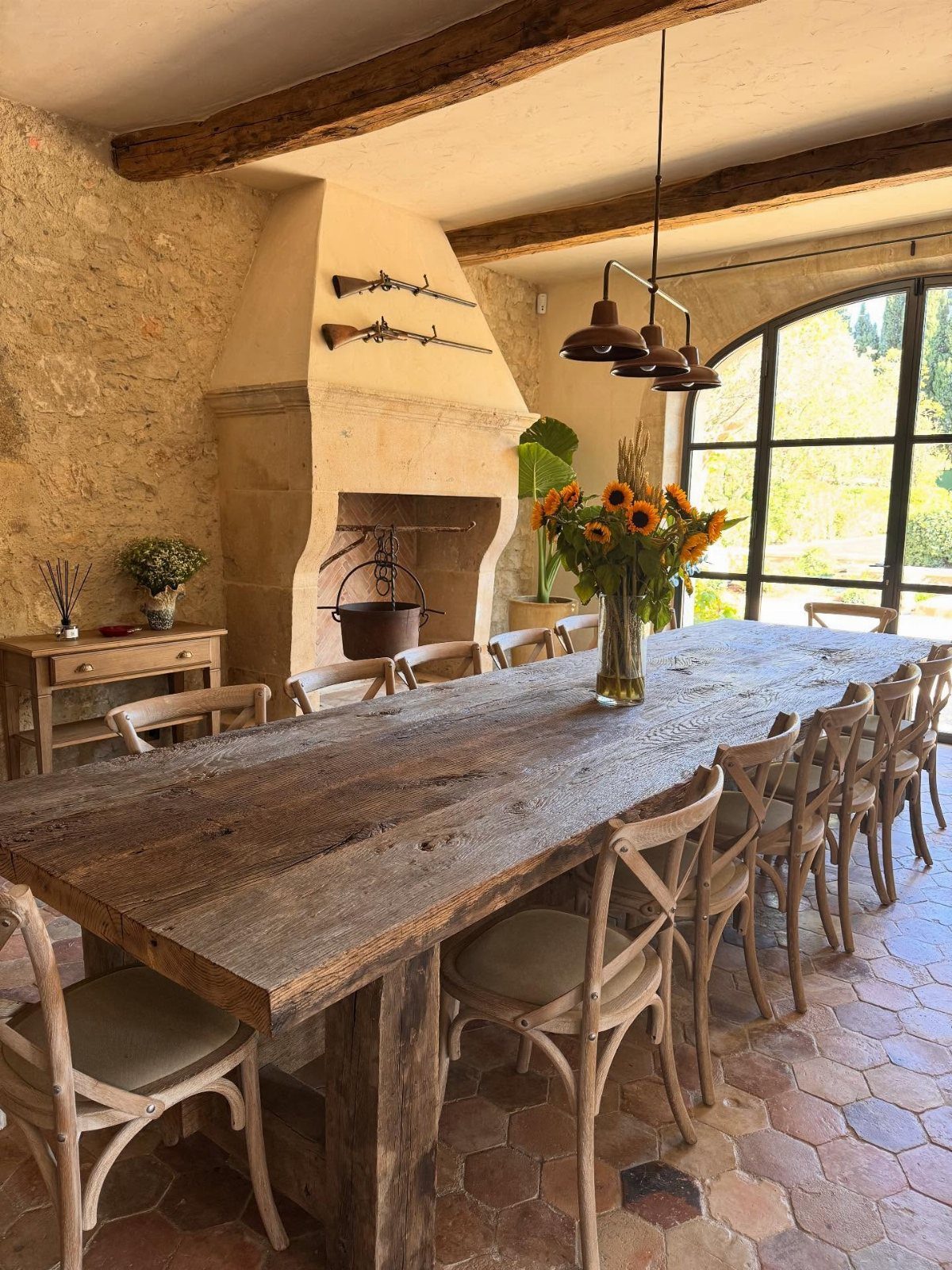 Rustic dining room with long wooden table, cross-back chairs, and terracotta tile flooring.