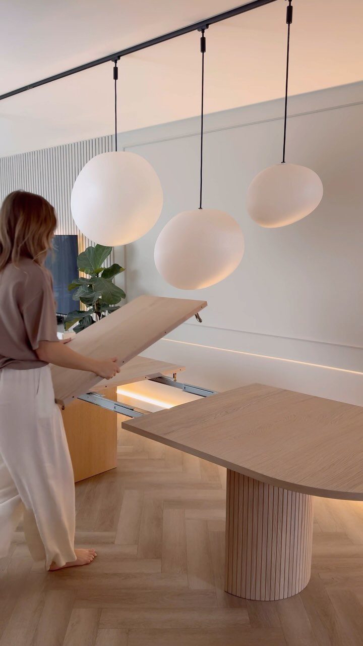 Woman extending a light wood dining table under three white spherical pendant lights.