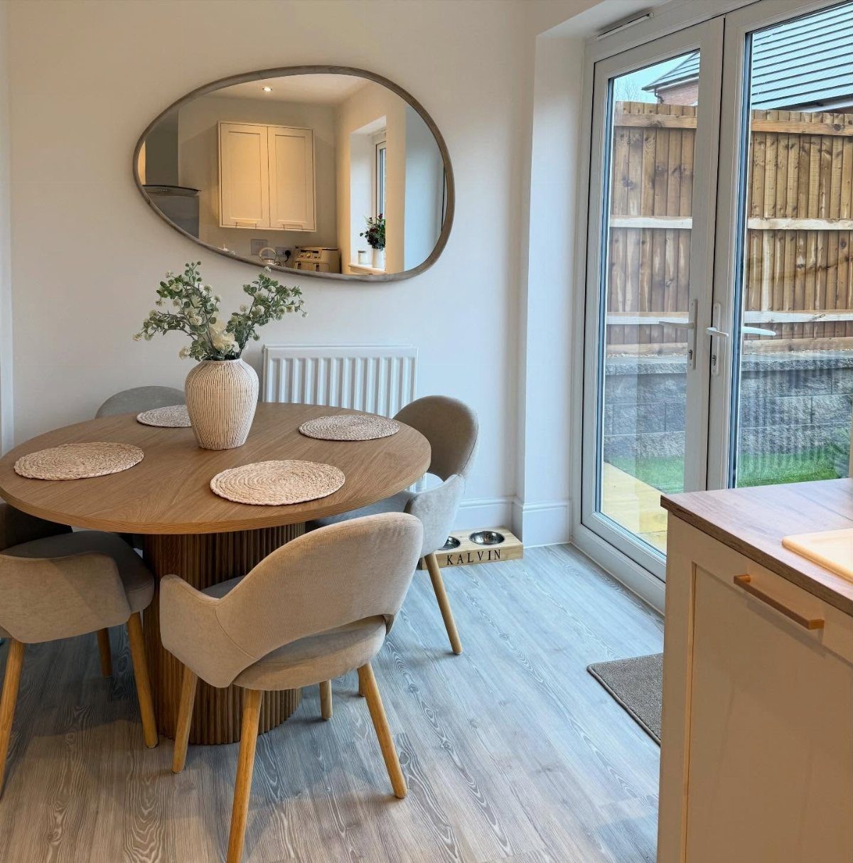 Round wooden dining table with beige chairs and an oval wall mirror.