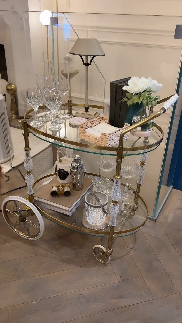 Gold bar cart with glass shelves holding glassware, flowers, and a small lamp.