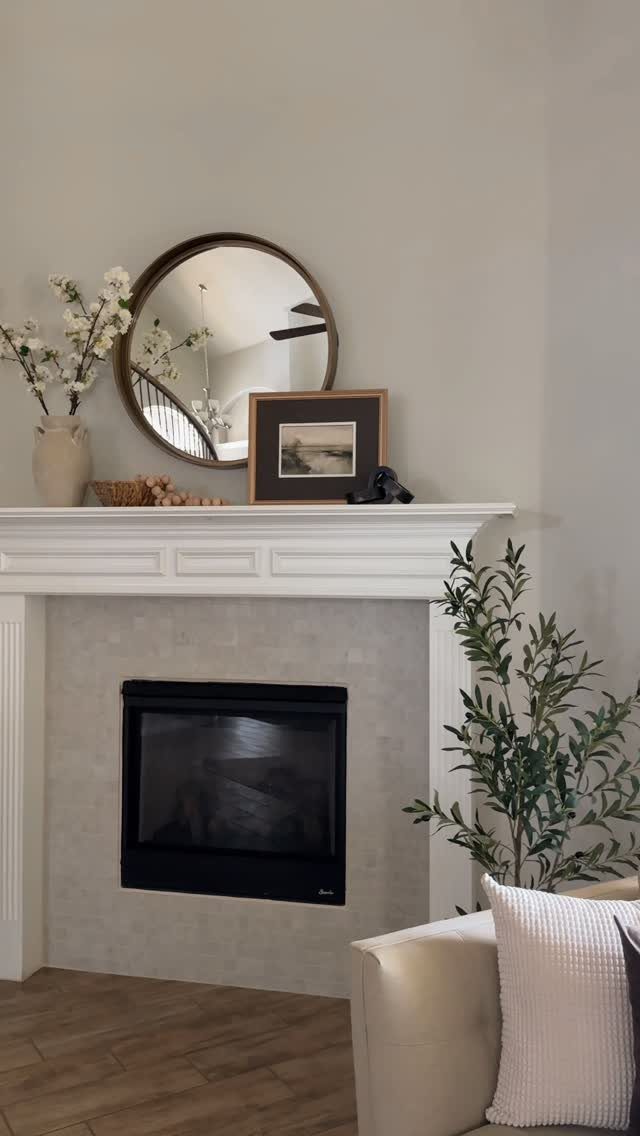 White fireplace mantel decorated with a round mirror, framed art, and white flowers.