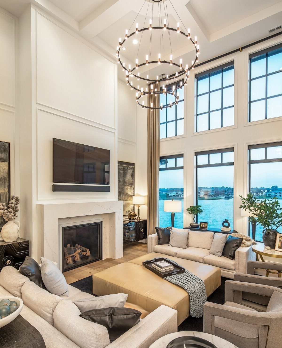 Modern living room with white fireplace mantel and large windows overlooking water.