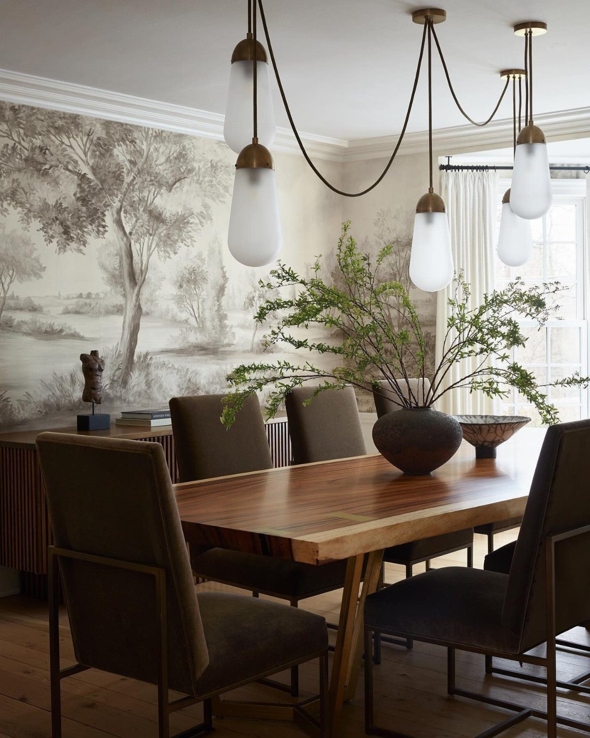 Wooden dining table with dark chairs and modern pendant lights against a landscape mural.
