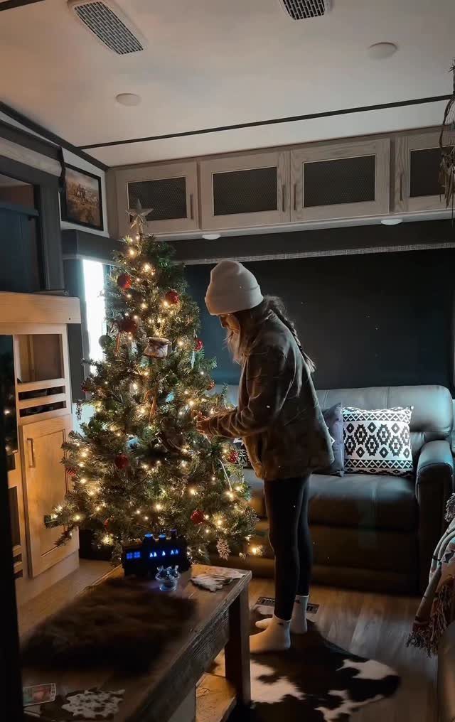 Woman decorating a lit Christmas tree inside a cozy mobile home living room