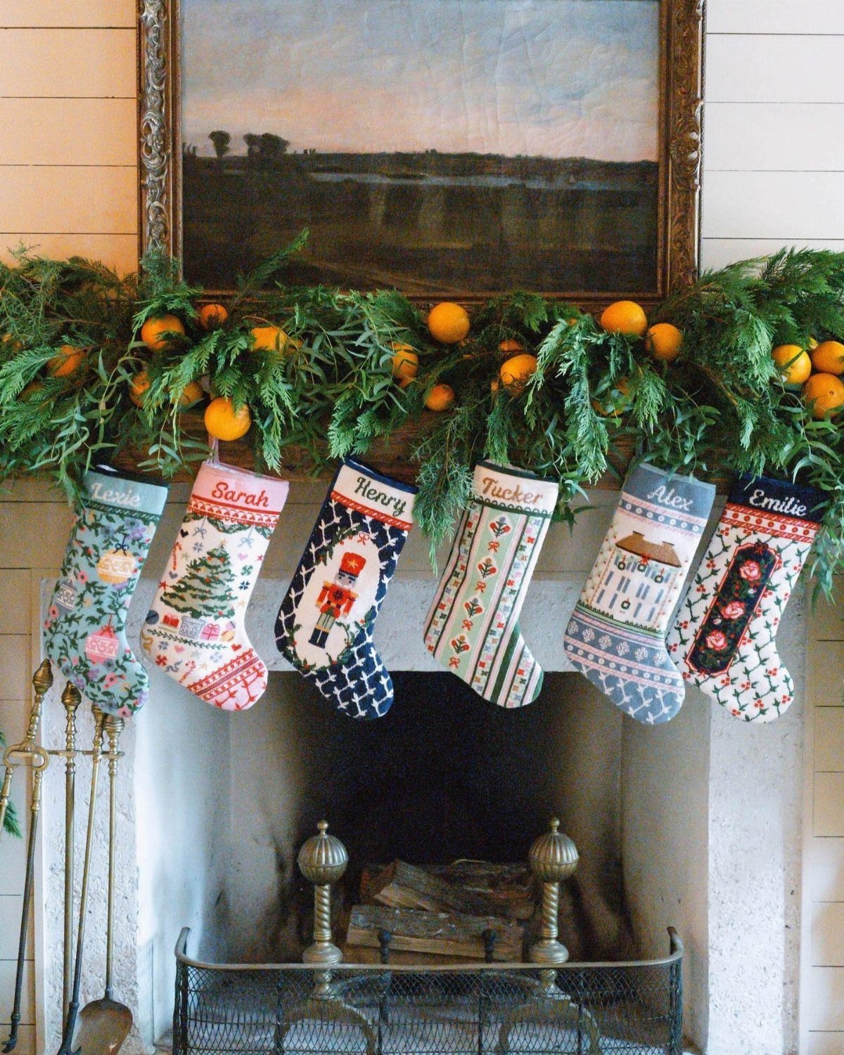 Six personalized patterned Christmas stockings hanging from a mantel with greenery and oranges.