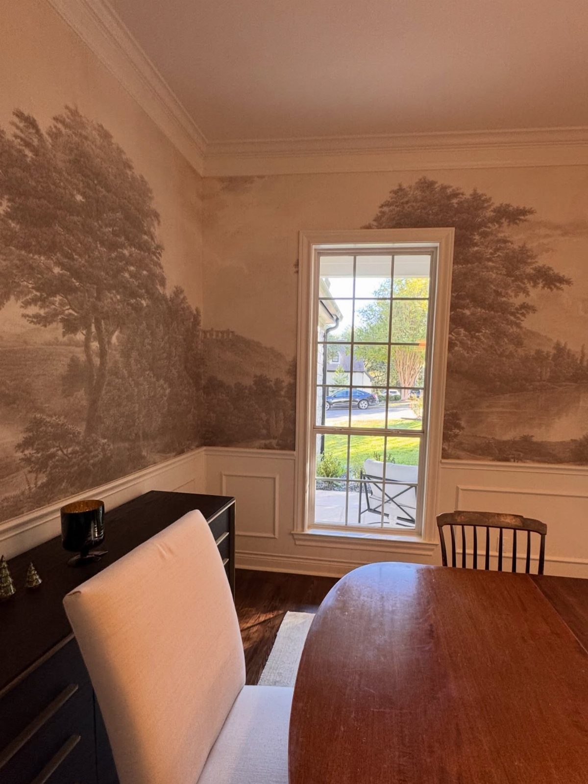 Sepia landscape wallpaper on dining room walls with white wainscoting and wooden furniture.