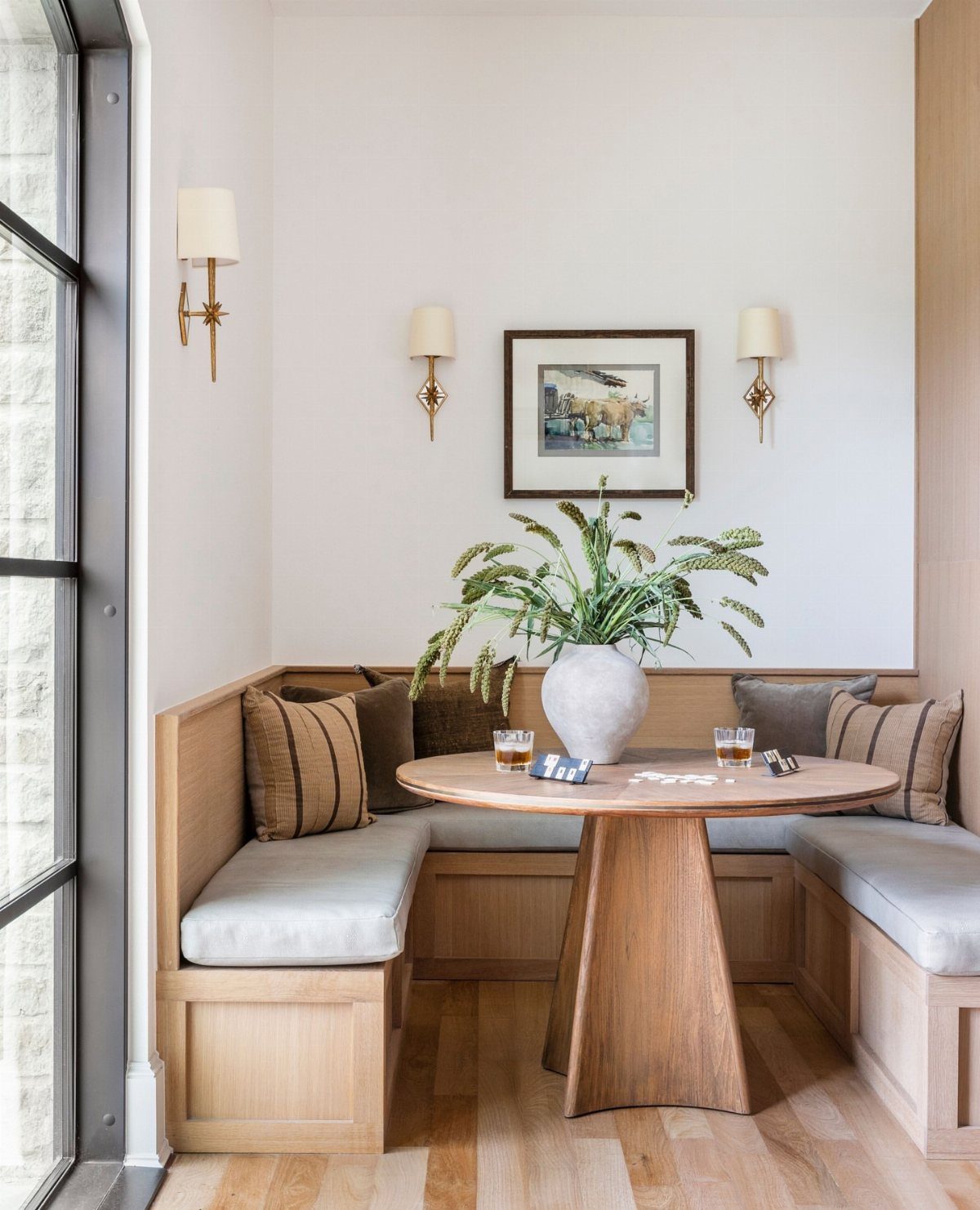 L-shaped wooden banquette with grey cushions and a round pedestal table in a nook.