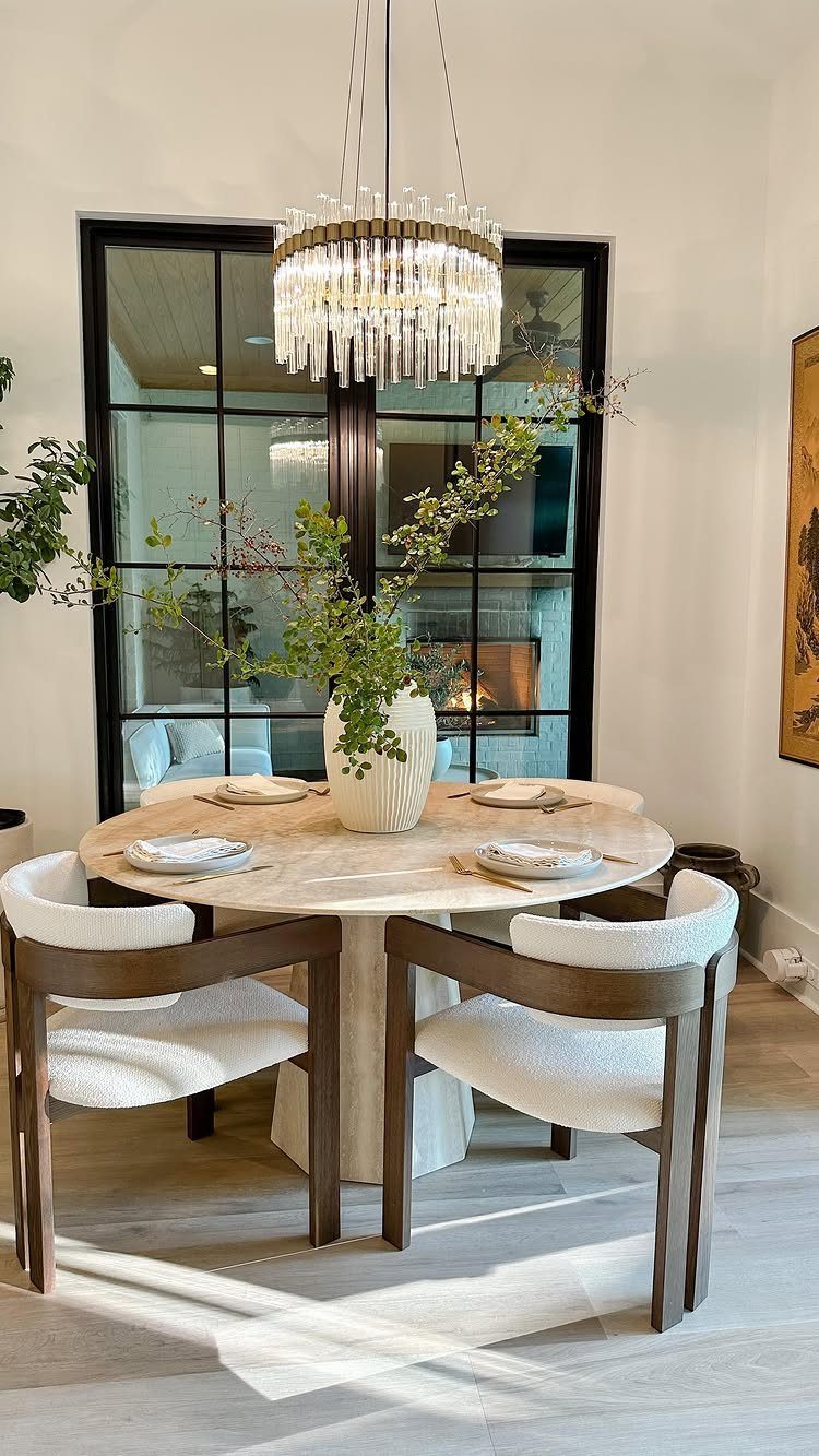 Round marble dining table with white upholstered chairs and green branches in a vase.