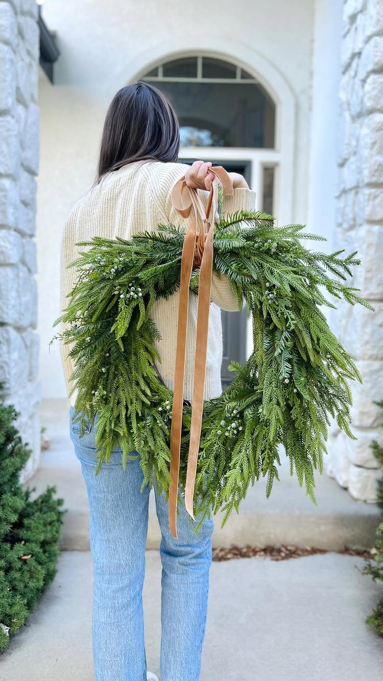 Woman holding a large green evergreen wreath tied with a long tan ribbon.