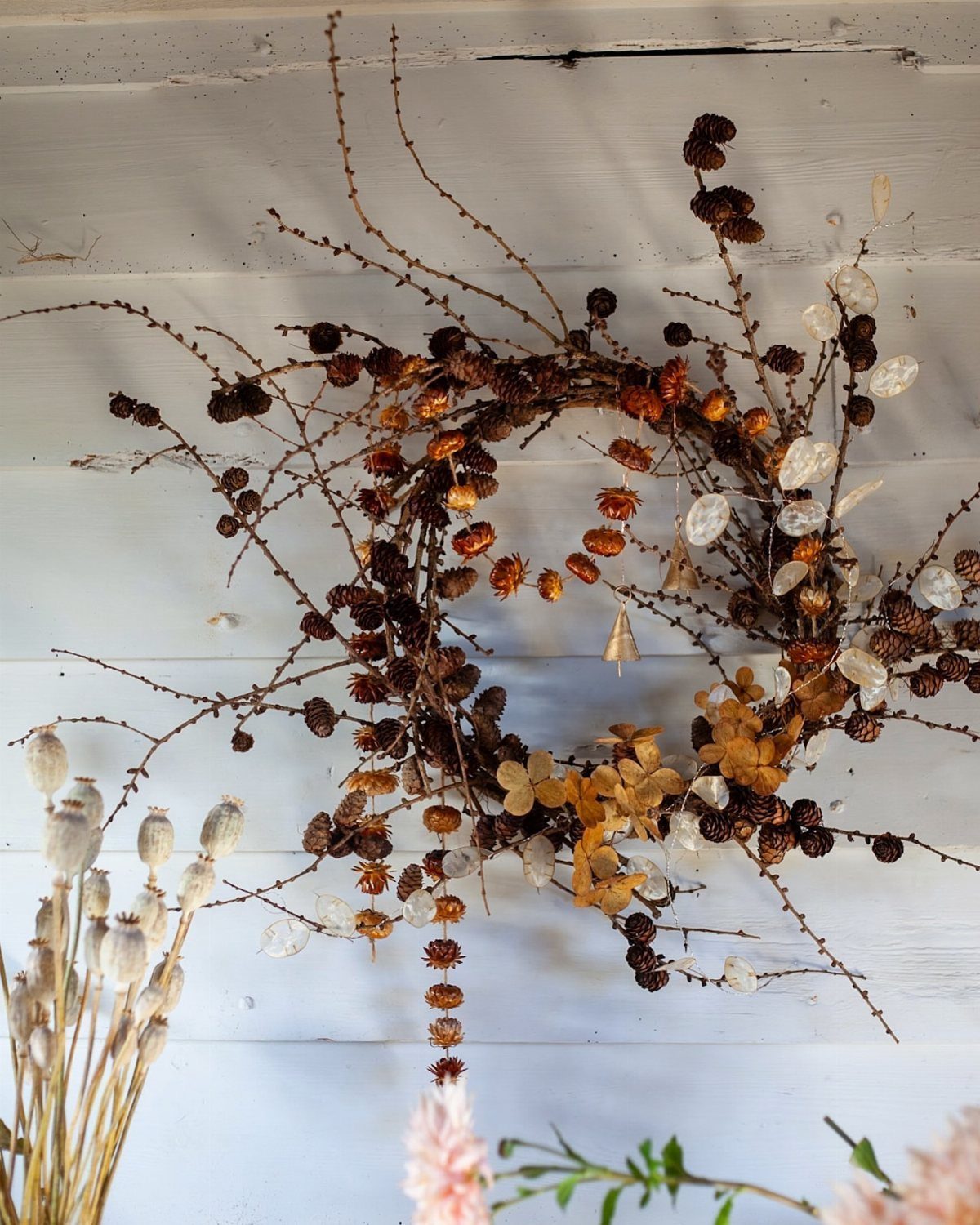 Rustic twig wreath with dried seed pods and orange botanicals on white wood.