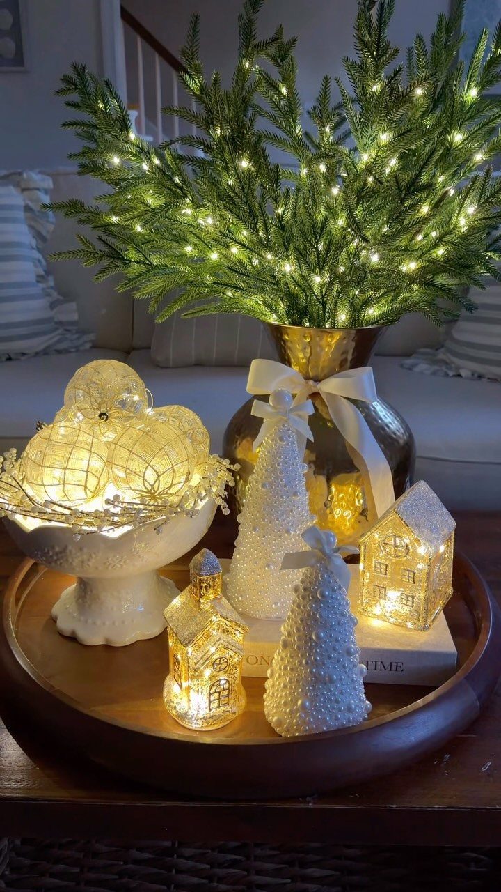 Glowing gold ornaments in a white bowl with miniature houses and lit greenery.