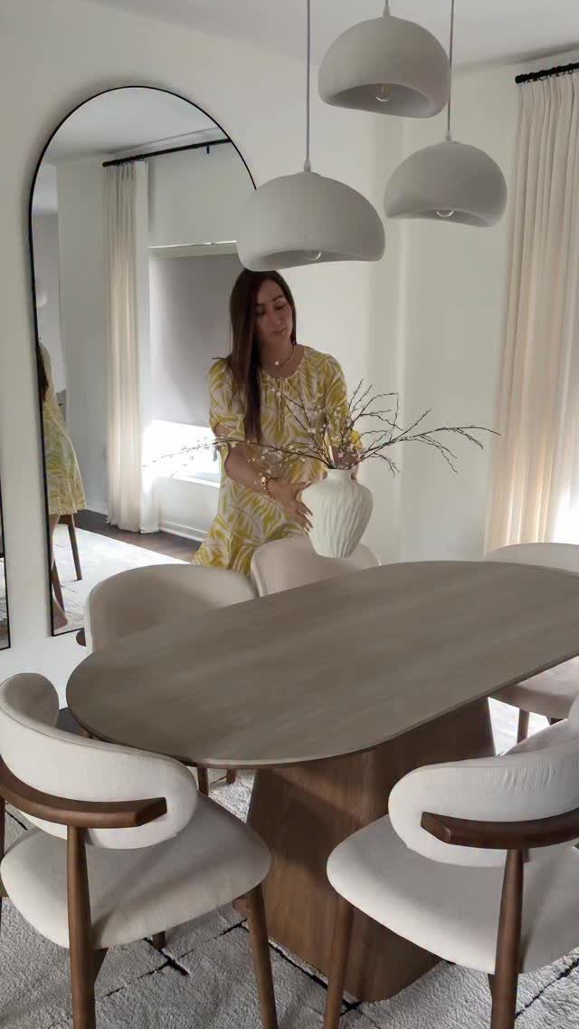 Woman standing by an oval wooden dining table with curved white chairs and pendant lights.