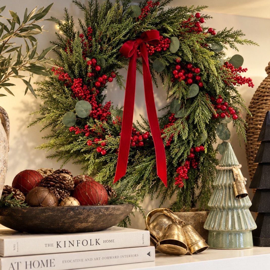 Green evergreen wreath with red berries and velvet bow above festive shelf decor