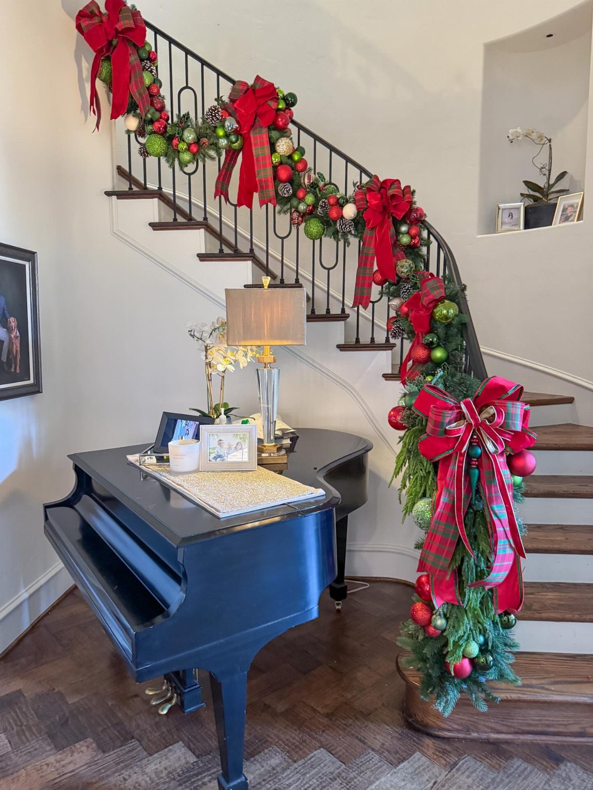 Staircase railing decorated with green garland, red plaid bows, and colorful ornaments