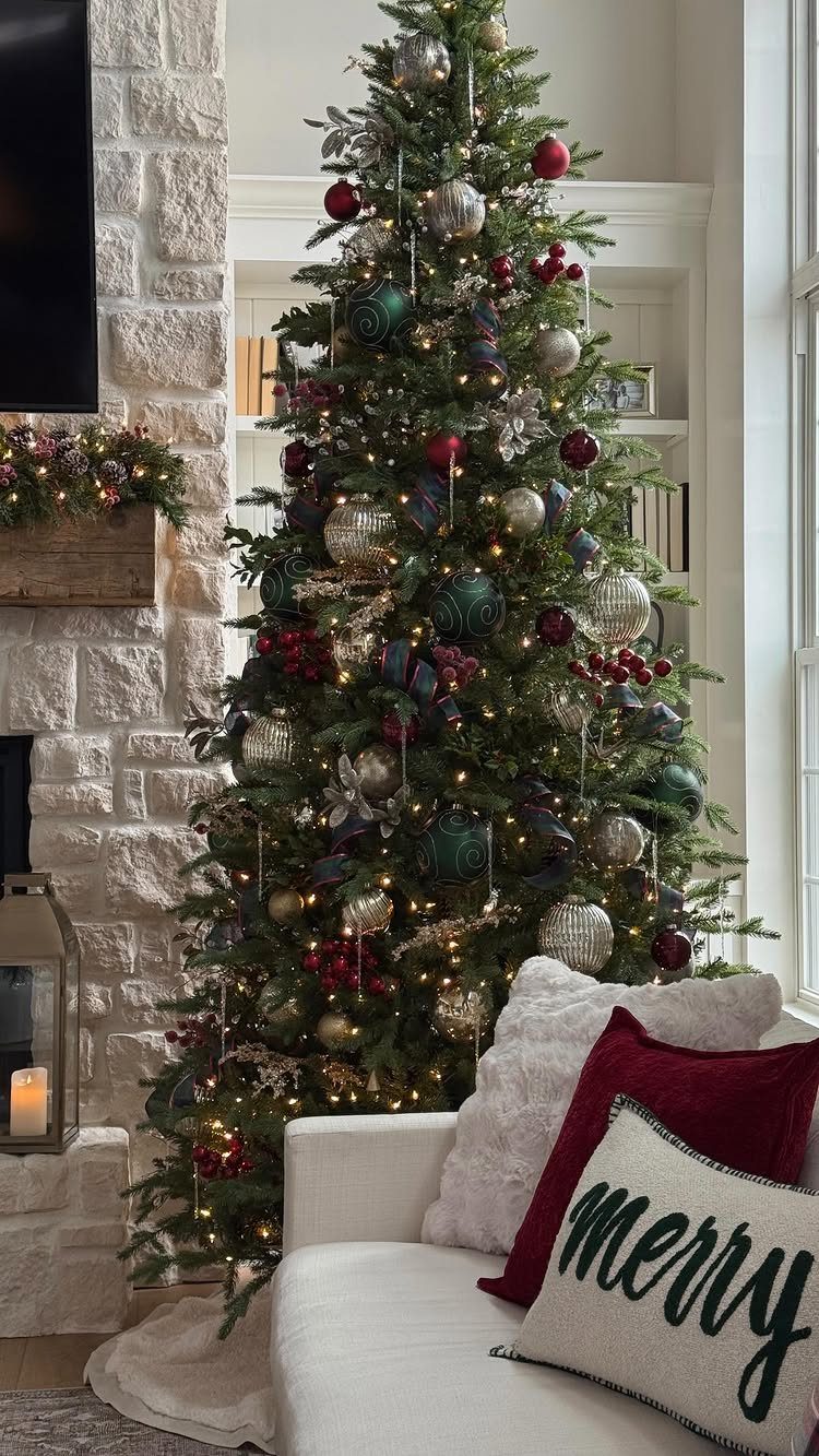 Decorated Christmas tree with red, green, and silver ornaments next to a stone fireplace.