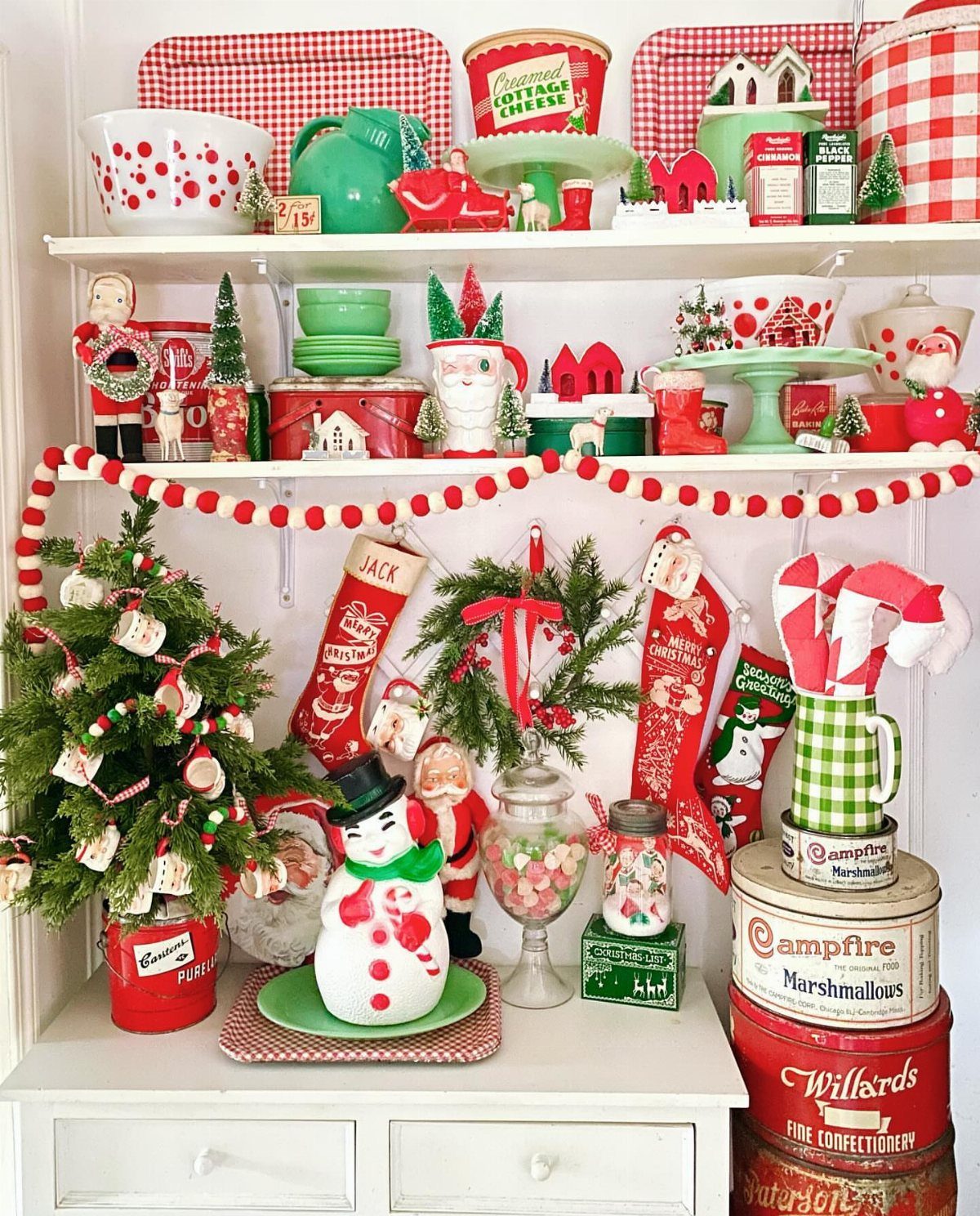 Vintage red and green Christmas collectibles arranged on white shelves with a small tree.