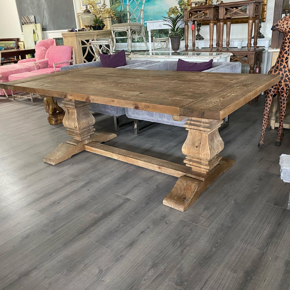 Natural wood trestle dining table with carved legs in a furniture showroom setting.