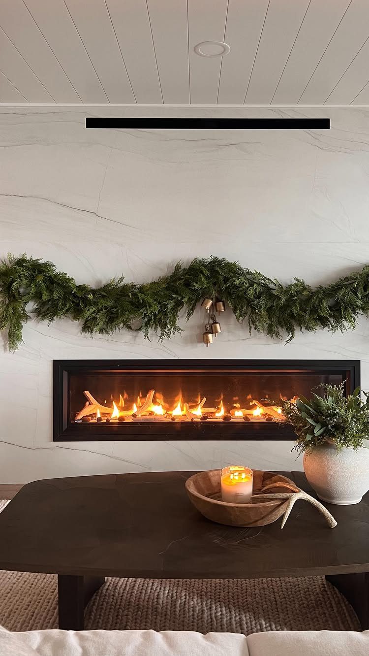 Modern white marble fireplace decorated with a green evergreen garland and lit fire.