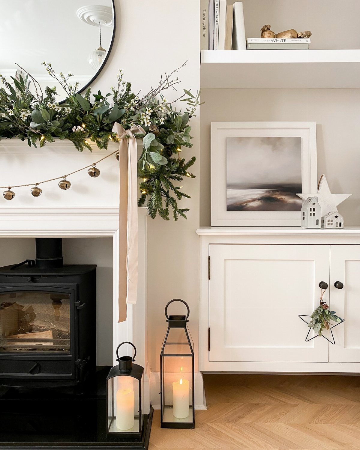 White fireplace mantel decorated with evergreen garland, gold bells, and lit floor lanterns.