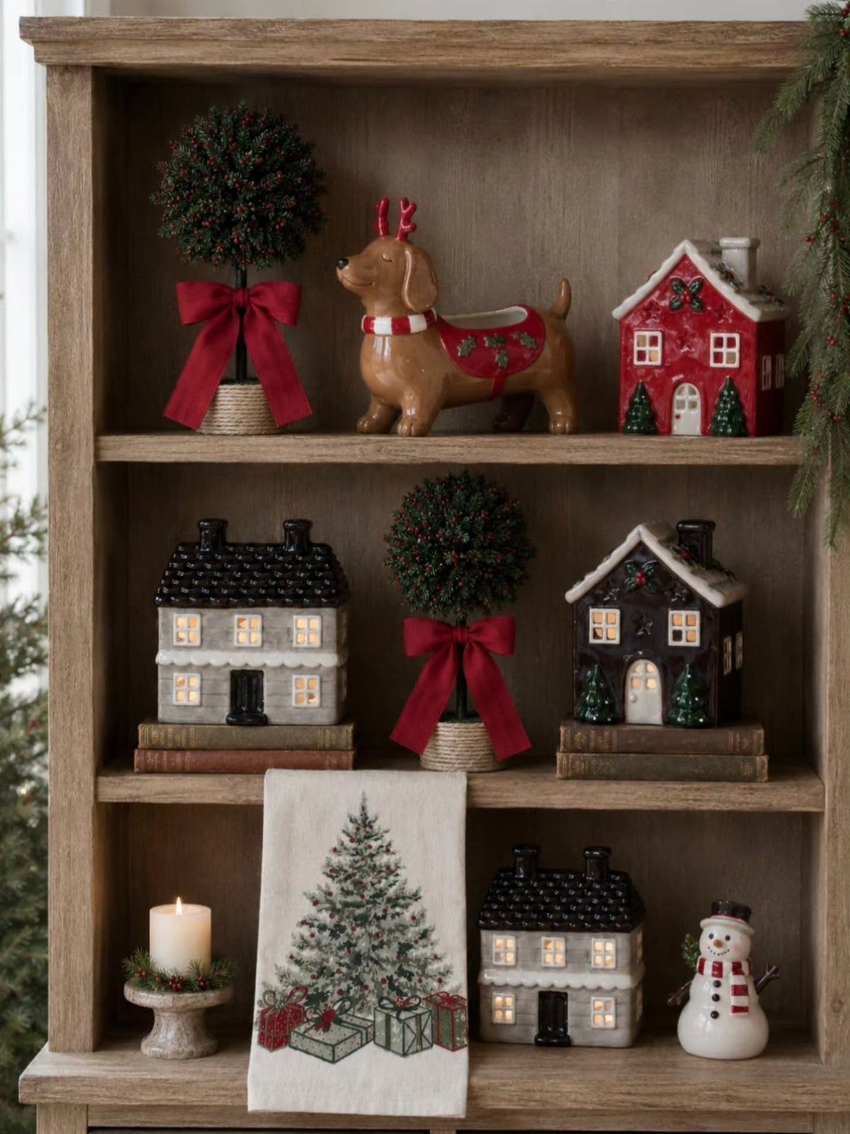 Wooden bookshelf styled with miniature Christmas houses, topiary trees, and holiday figurines.