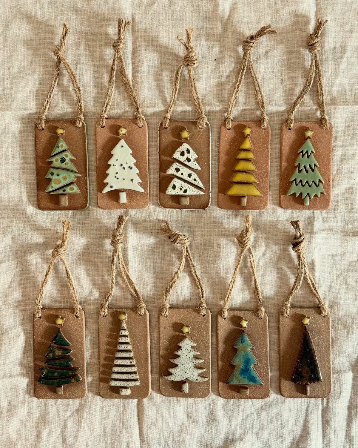 Ten handmade clay Christmas ornaments with colorful tree designs on a white fabric.