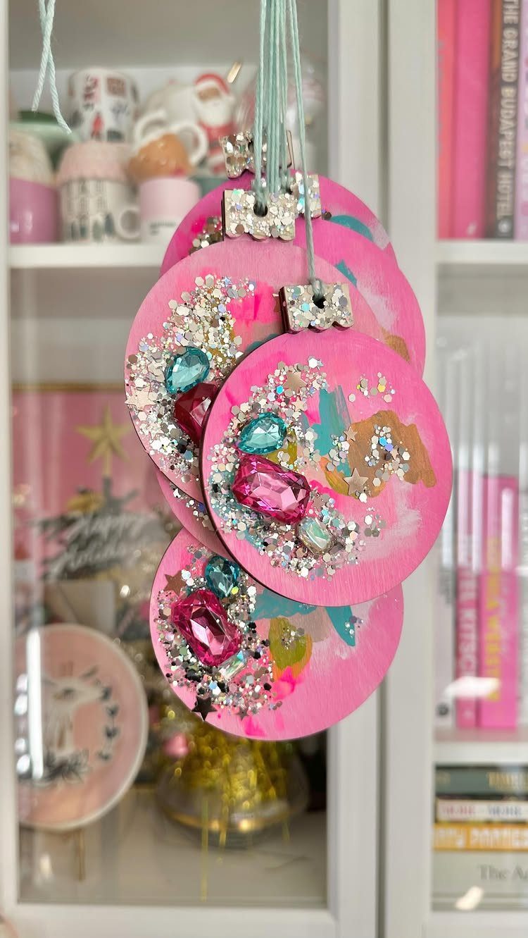 Pink round ornaments decorated with colorful glitter and large faux gemstones hanging by strings.