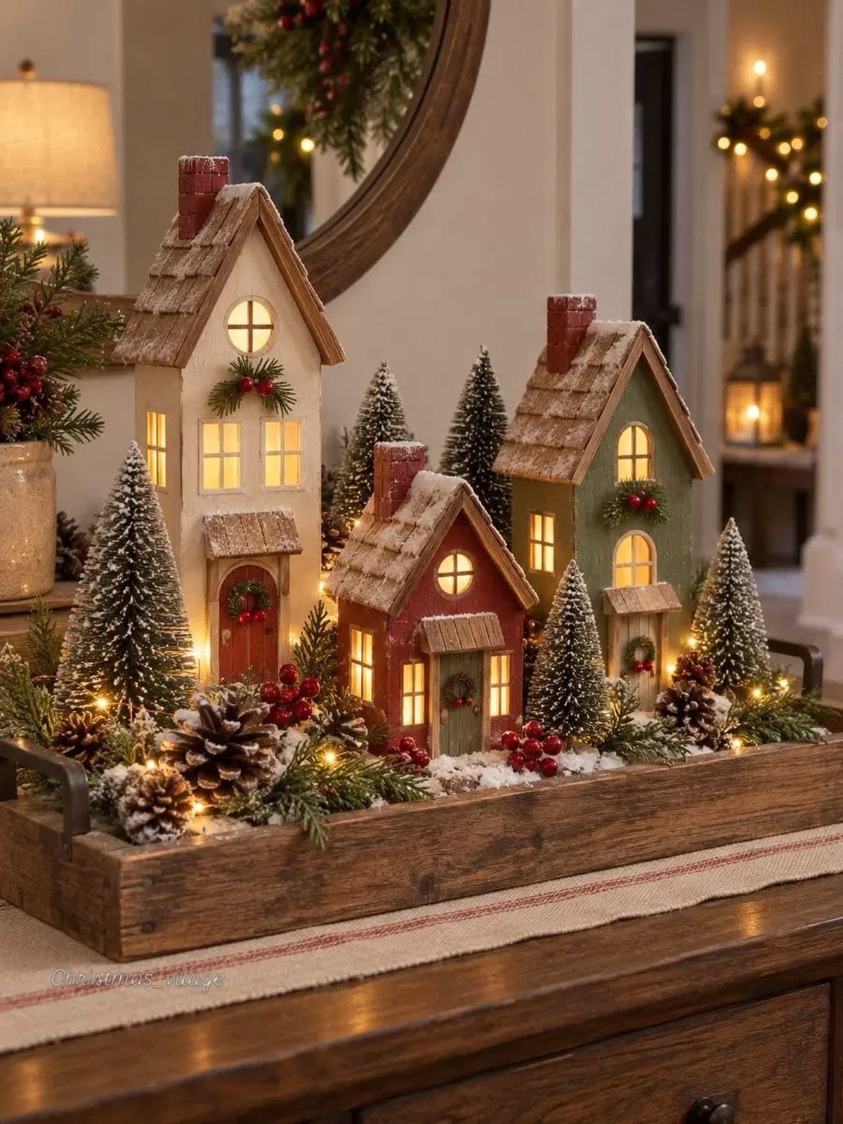 Miniature wooden Christmas houses with lights and snow in a rustic wooden tray.