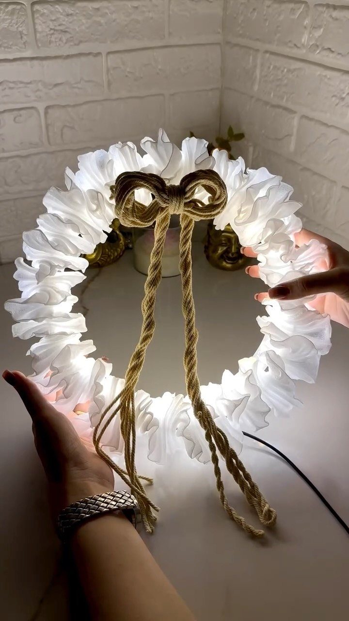 White ruffled fabric wreath with internal lights and a brown twine bow.