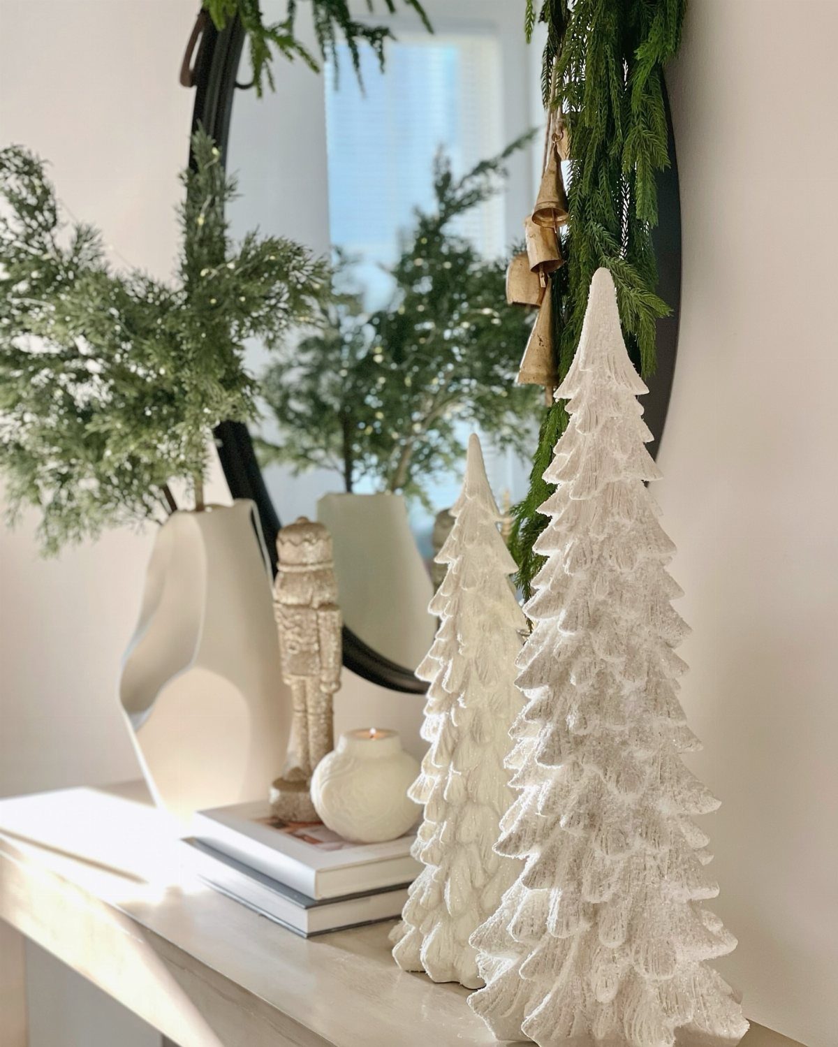 White decorative Christmas trees and greenery on a light-colored console table with mirror.