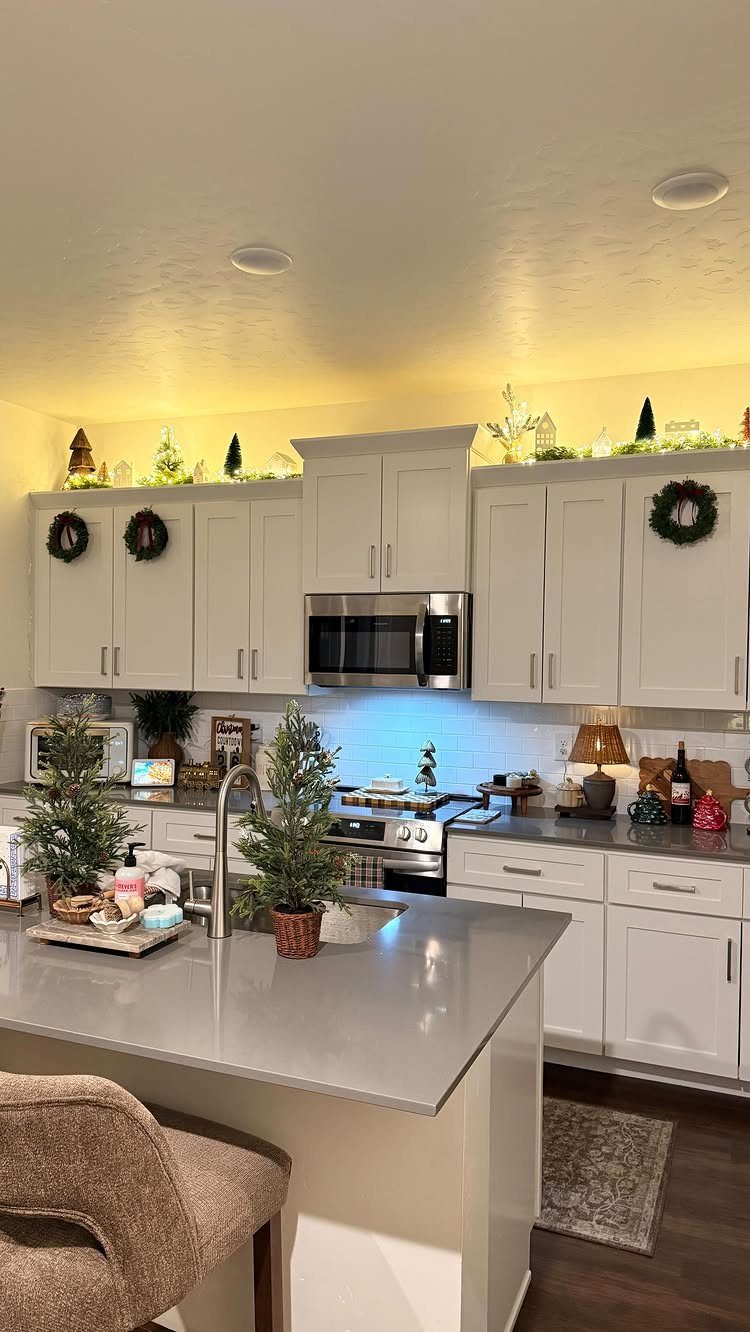 Kitchen decorated for Christmas with small trees, wreaths, and lit cabinet top greenery.