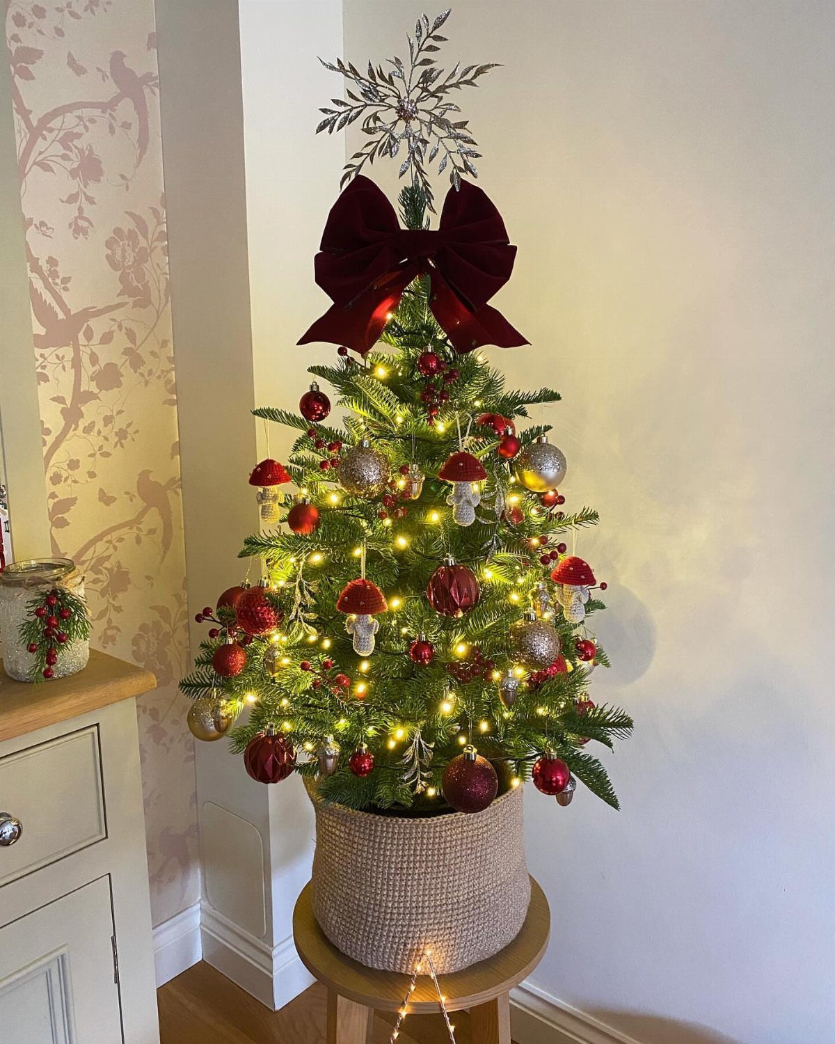 Small decorated Christmas tree in a woven basket on a wooden stool.