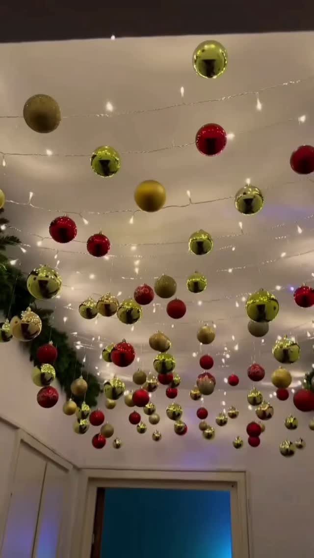 Red, gold, and green Christmas ornaments hanging from string lights on a ceiling