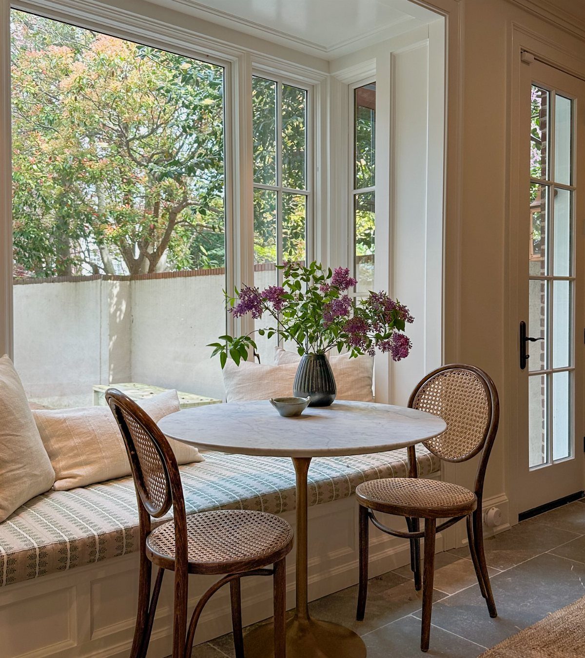 Round white marble cafe table with two woven chairs in a bright sunroom.