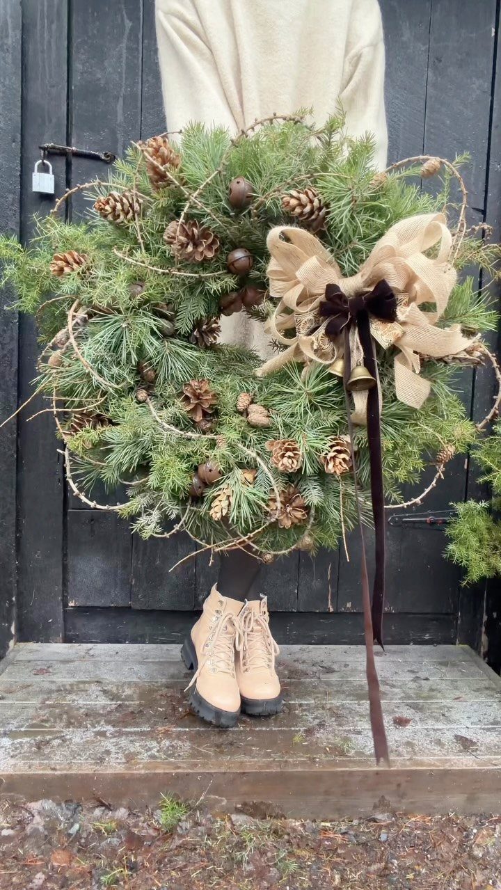 Evergreen wreath with pinecones and a burlap bow held against a black door.