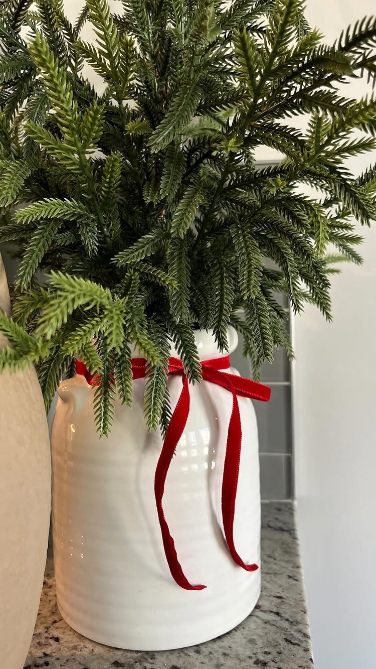 Evergreen branches in a white ceramic pitcher tied with a red velvet ribbon.